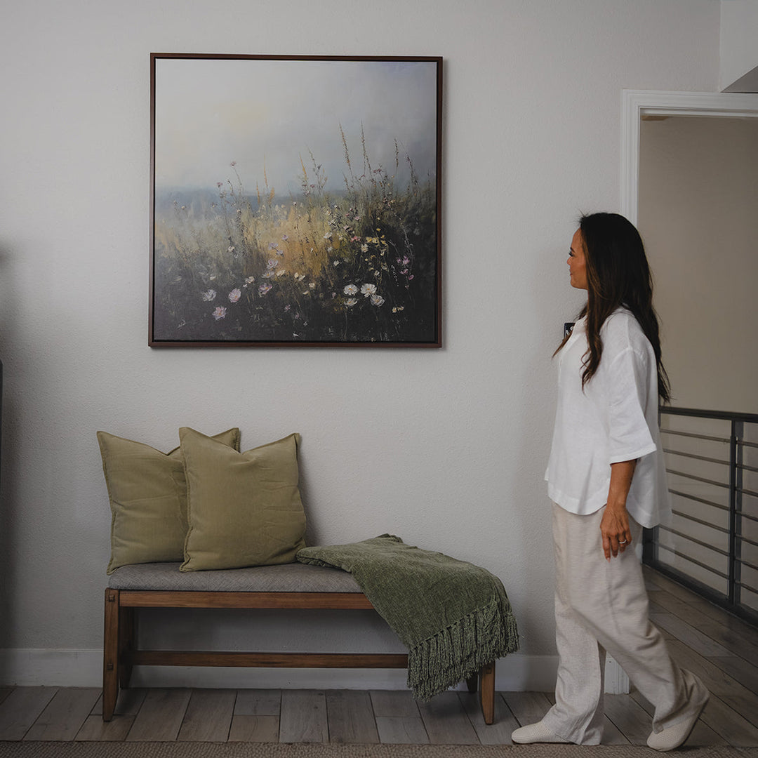 A cozy living room with a white sectional sofa, assorted neutral and blue pillows, a rustic coffee table, and a framed landscape painting of wildflowers on a white shiplap wall.