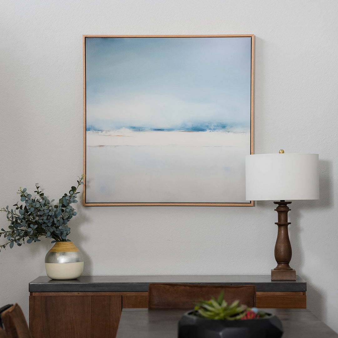 A neutral-toned living room with a beige sofa, light cushions, a wooden side table with a cream lamp, dried flowers in a vase, and a large abstract painting in soft blue and white hues on the wall.