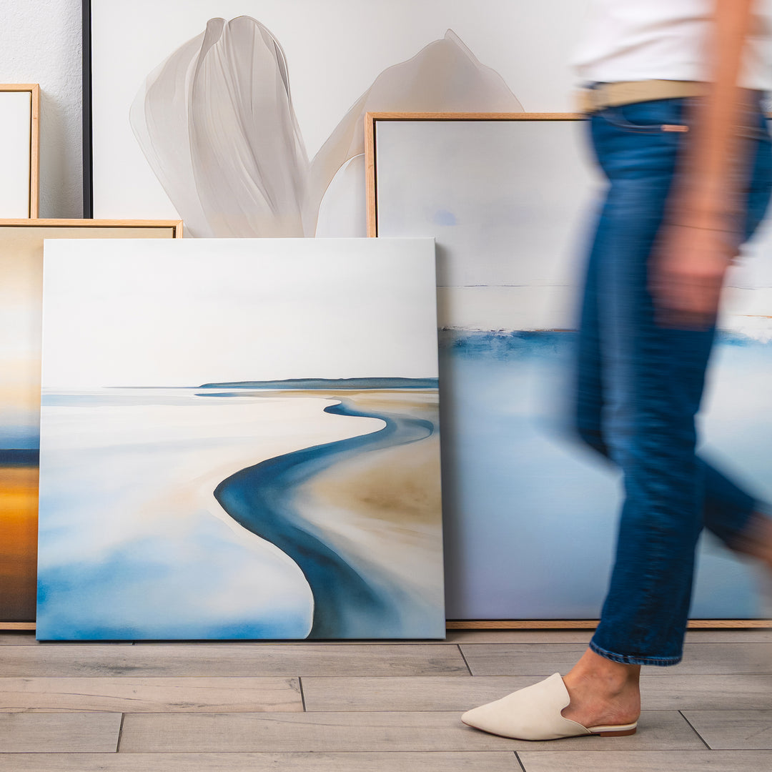 A person in jeans and white shoes walks past three Serene Coastline Canvas Art pieces with soft, muted tones leaning against a wall on a wooden floor, adding a calm vibe to modern interiors.
