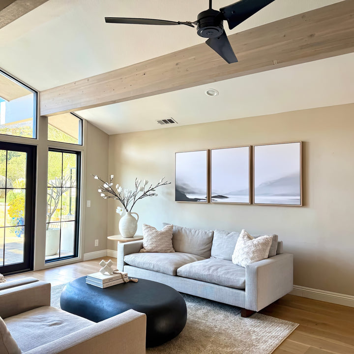 A modern living room with a beige sofa, light armchair, and black oval coffee table features the Serenity Peaks Set of 3 minimalist landscape art on the wall. Large windows and neutral decor invite natural light, while a vase with branches adds an organic touch.