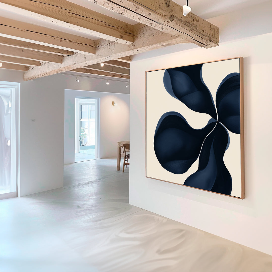 A modern, minimalist room with exposed wooden beams and white walls features Blossom Blue Abstract Canvas Art—a large print with a bold, dark blue abstract design. Natural light enhances this striking minimalist wall art.