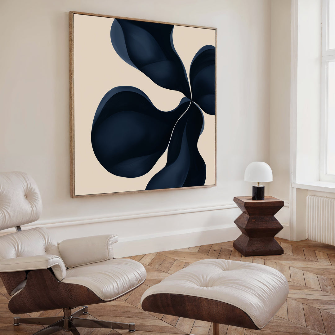 A modern living room with a cream lounge chair and ottoman, dark wooden side table, white lamp, and Blossom Blue Abstract Canvas Art featuring navy and beige above the seating area.