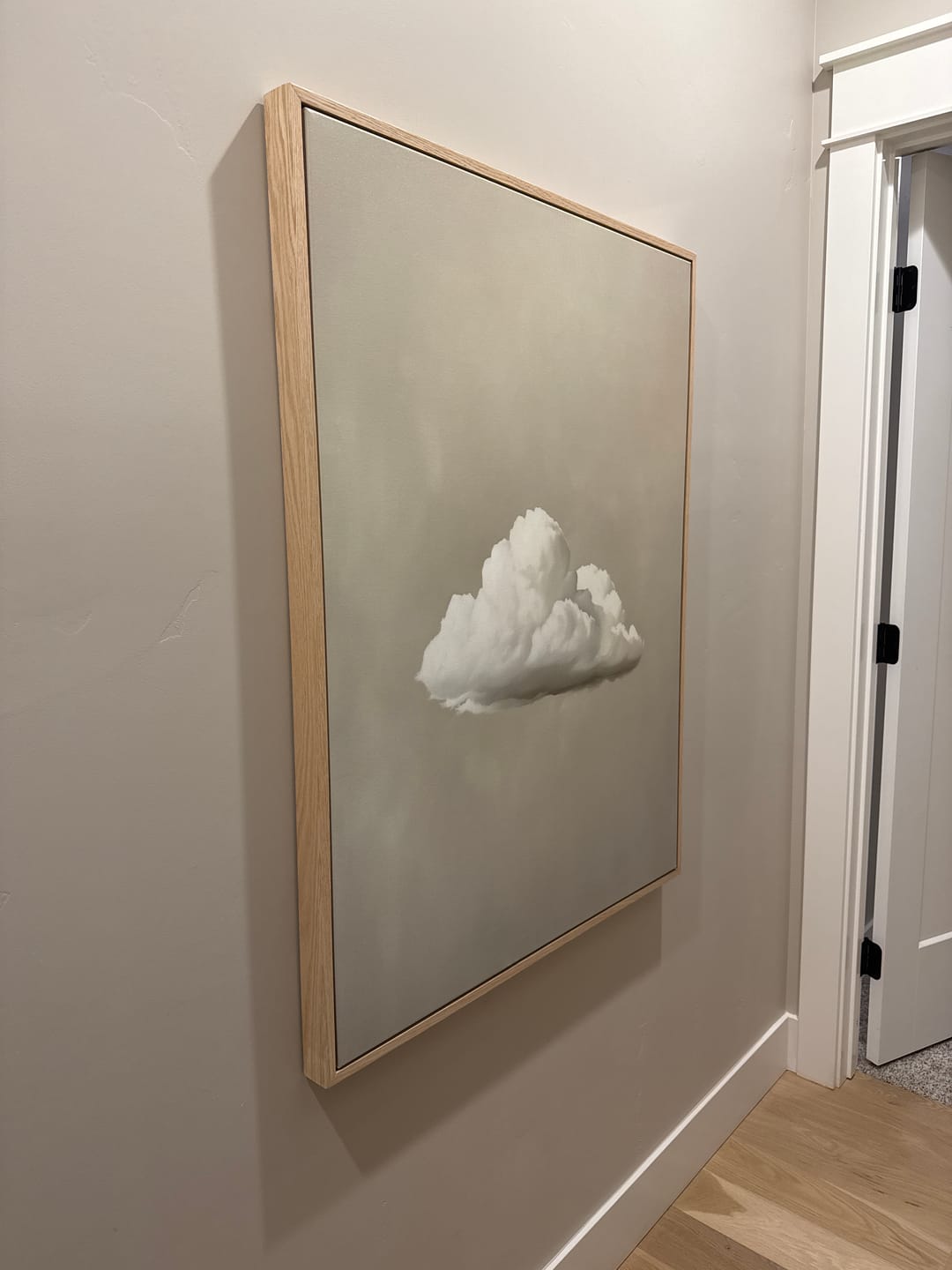 The Morning Sky Abstract Canvas Art, featuring a single white cloud on a soft gray background, hangs on a beige hallway wall near a white doorframe, bringing minimalist style and serene decor to the space.