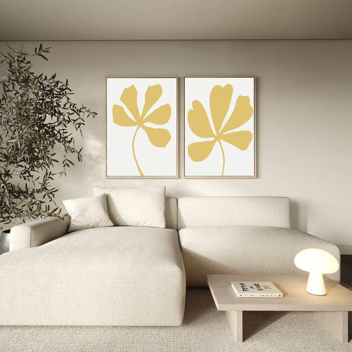 A minimalist living room with a light beige sectional sofa, a small wooden table with a glowing lamp and books, a leafy plant, and the Sunleaf Silhouettes Set of 2 floral wall art prints above the sofa.