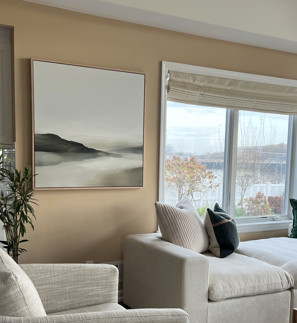 A cozy living room with a beige wall features Soft Valley Canvas Art, light gray armchairs, a sectional sofa with neutral and green pillows, a potted plant, and a window overlooking trees and water.