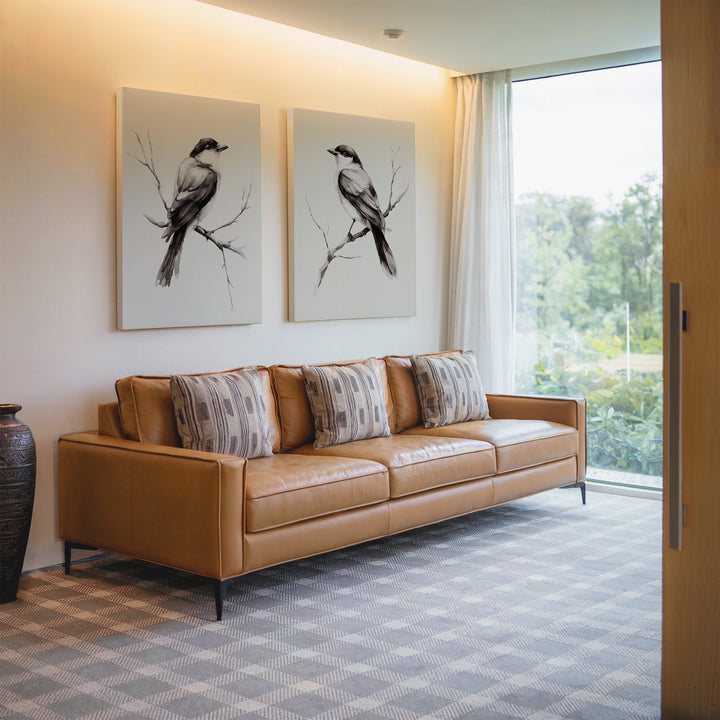 A modern living room with a tan leather sofa, patterned cushions, Still Perch Set of 2 bird art prints on the wall, a decorative vase, and large windows overlooking Nordic Park’s lush greenery.