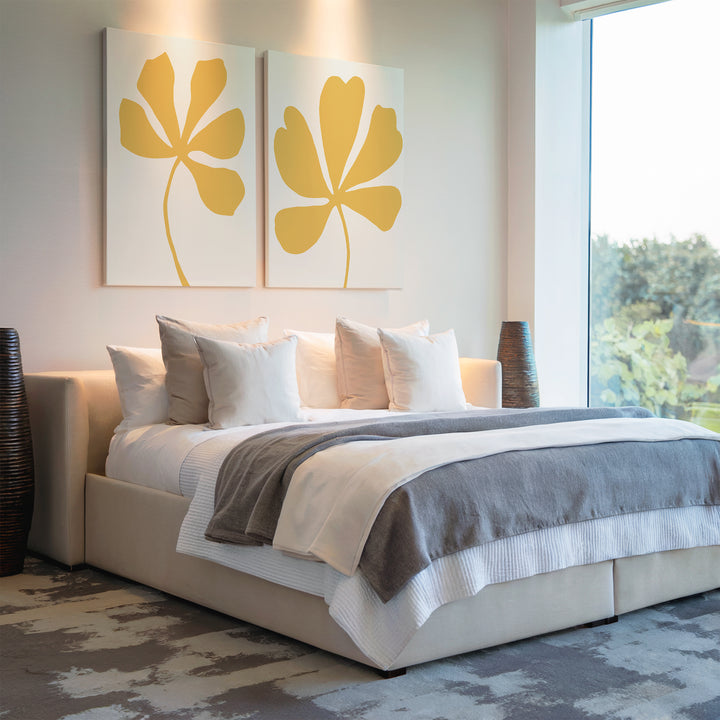 A modern bedroom with a neatly made bed in white and gray, multiple pillows, and two Sunleaf Silhouettes Set of 2 yellow floral wall art above the headboard, complemented by large windows letting in natural light.