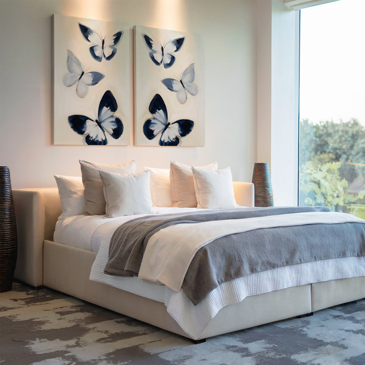 Modern bedroom with a large bed in white and gray bedding, several white pillows, two wicker lamps, and Midnight Wings Set of 2 butterfly wall art above the headboard. A large window on the right fills the space with natural light.