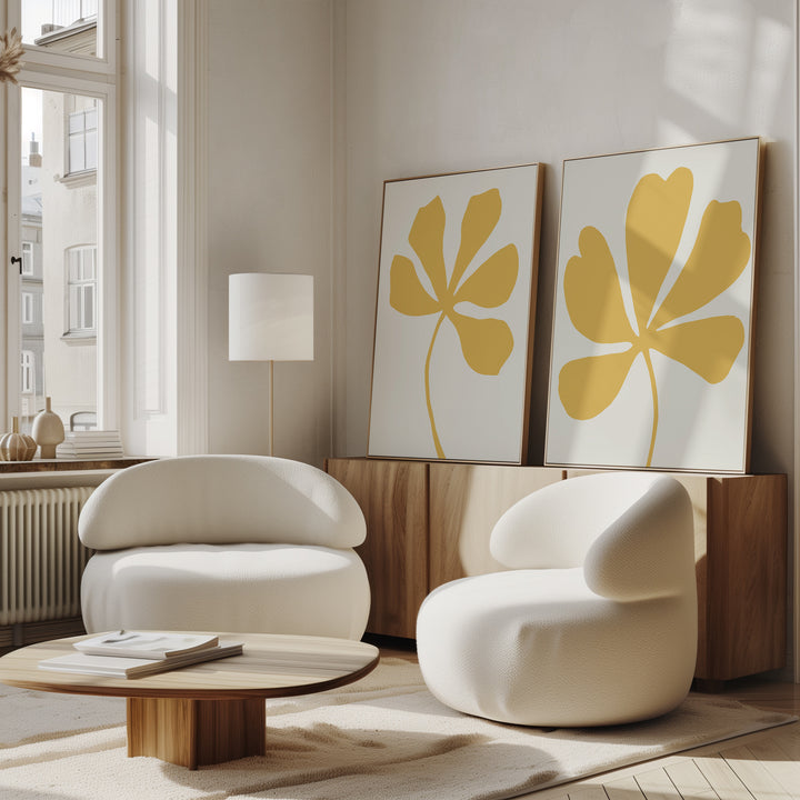 A modern living room with two white rounded chairs, a low wooden coffee table, and the Sunleaf Silhouettes Set of 2 wall art prints featuring yellow leaf shapes, set against light walls and large windows letting in natural light.