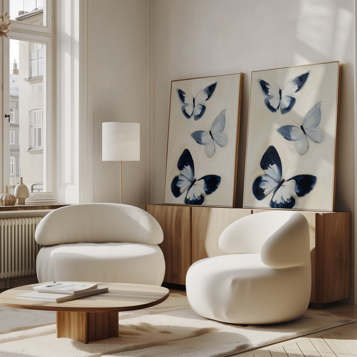 Modern living room with two curved white chairs, a round wooden coffee table, and the Midnight Wings Set of 2 butterfly wall art in blue and white on a wooden sideboard beside a tall window with natural light.