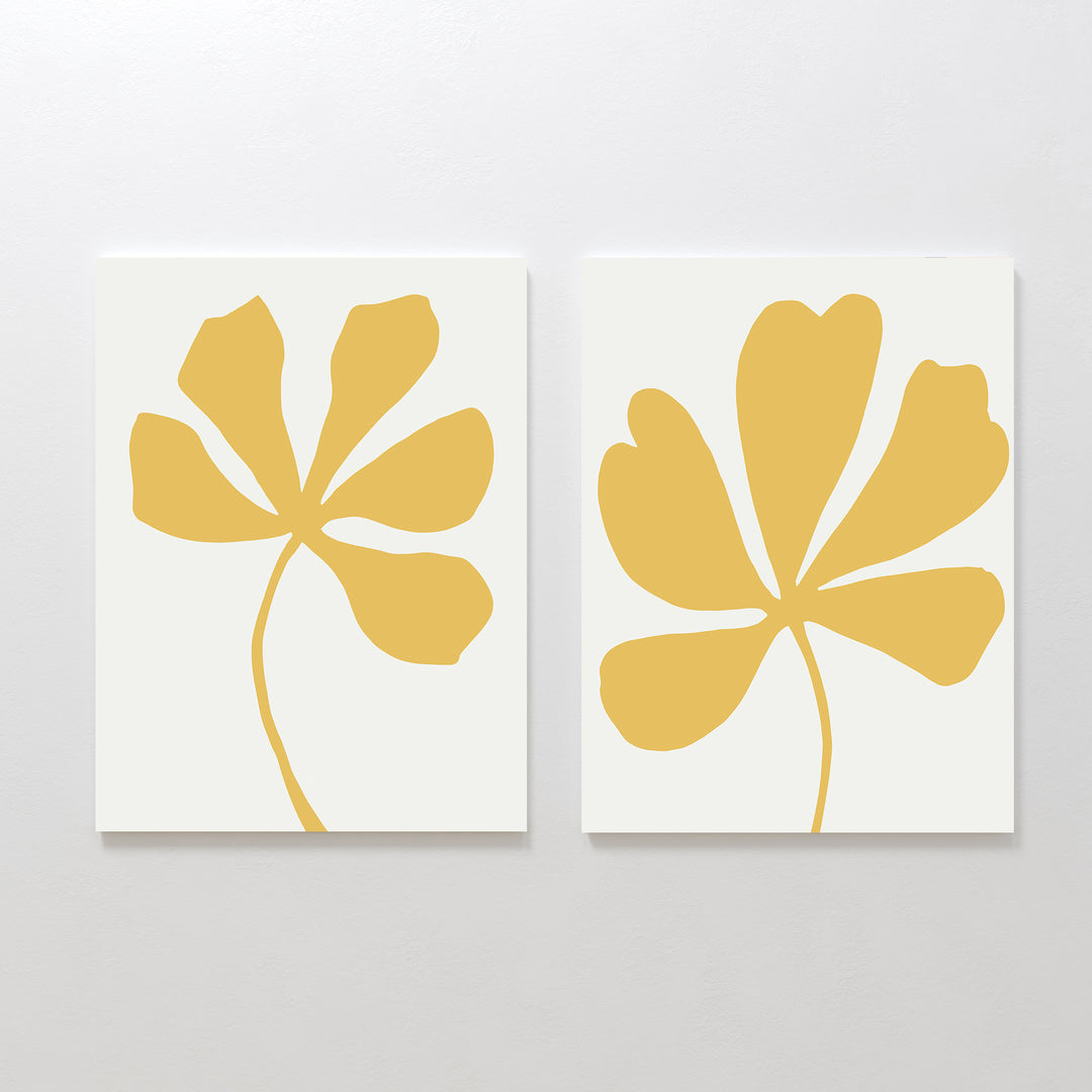 The Sunleaf Silhouettes Set of 2 features abstract golden-yellow flower shapes with curving stems on white backgrounds, creating a modern floral design. Displayed side by side, they add a minimalist touch to any light-colored wall.
