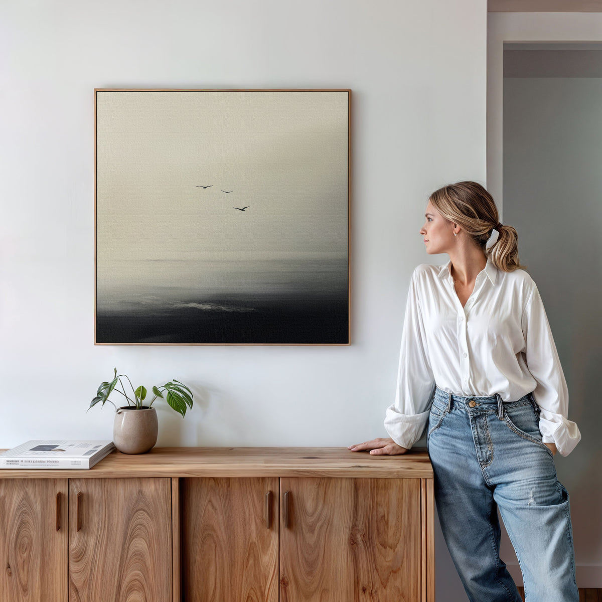 A woman in a white blouse and jeans stands by a wooden cabinet, admiring the Above the Calm Canvas Art—a large framed painting of birds over misty water—paired with a potted plant and books for a serene bedroom look.