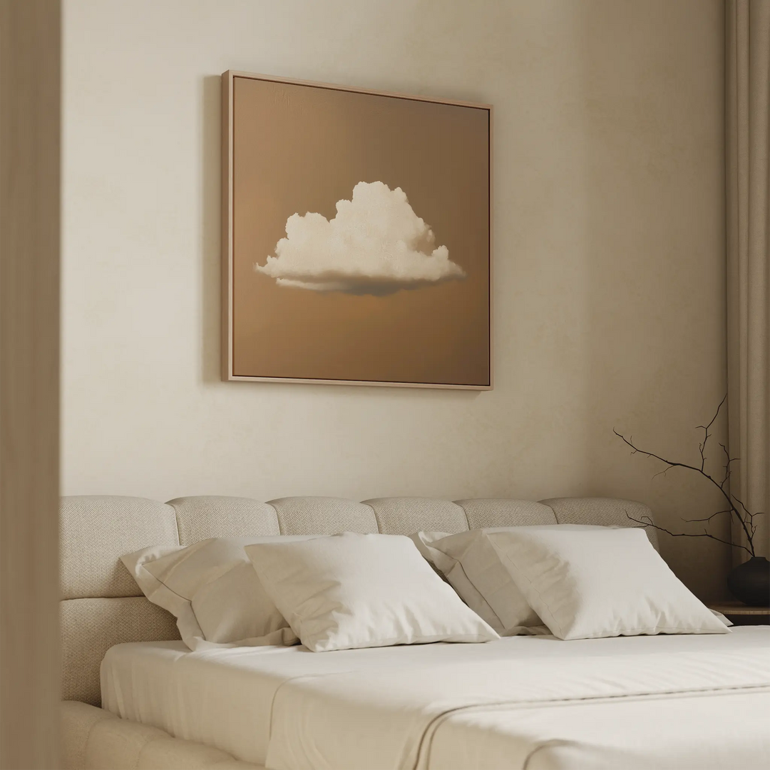 A minimalist bedroom features a beige upholstered bed, white pillows, and a tan wall. Above the bed is the Amber Sky Cloud Canvas Art—a modern framed picture of a white cloud on brown—enhancing the serene decor.