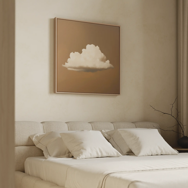 A minimalist bedroom features a beige upholstered bed, white pillows, and a tan wall. Above the bed is the Amber Sky Cloud Canvas Art—a modern framed picture of a white cloud on brown—enhancing the serene decor.