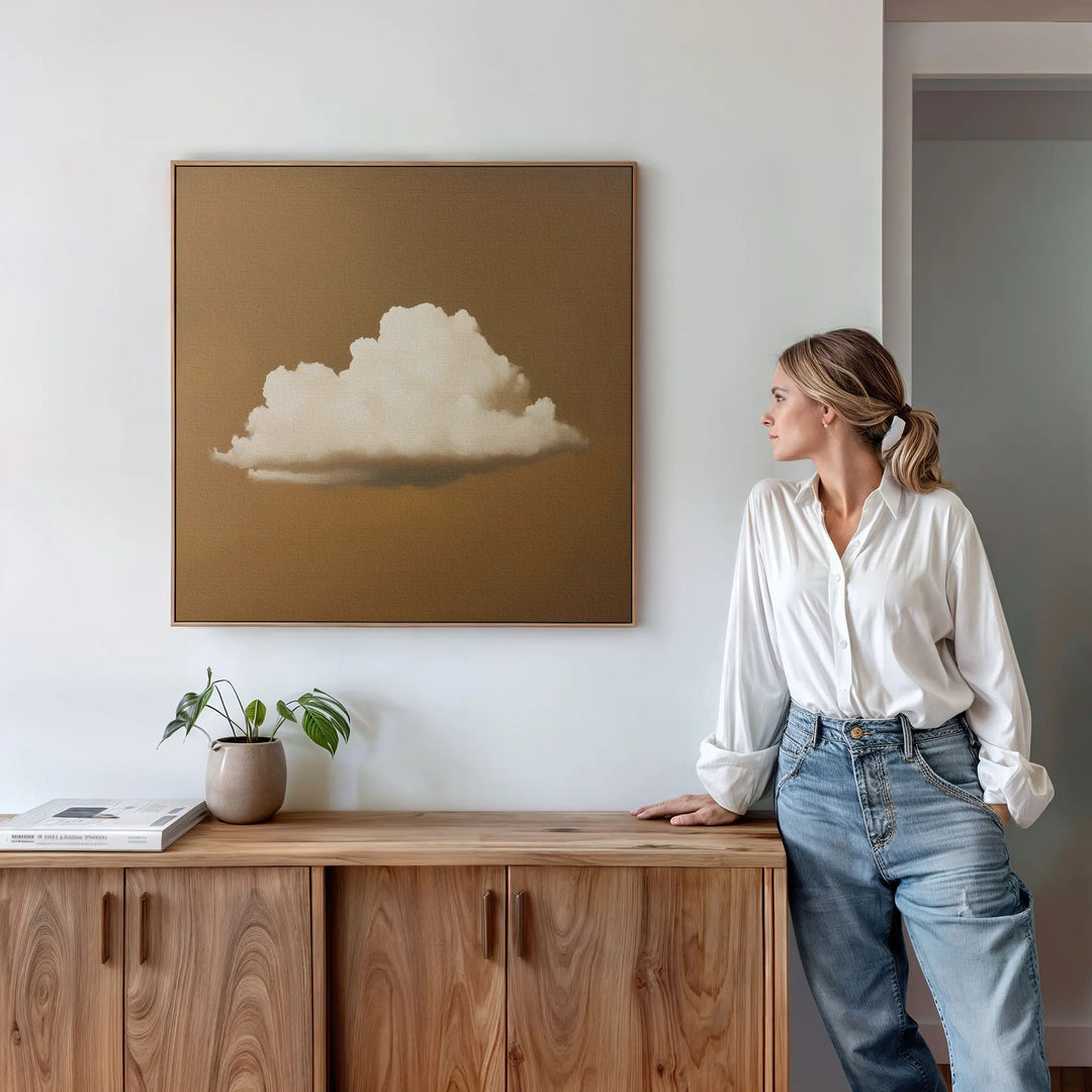 A woman in a white blouse and jeans leans against a wooden sideboard, admiring the Amber Sky Cloud Canvas Art—a minimalist cloud painting on brown—hanging on a white wall. A potted plant and books complete the stylish bedroom decor.