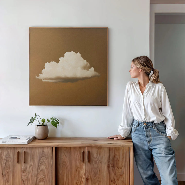 A woman in a white blouse and jeans leans against a wooden sideboard, admiring the Amber Sky Cloud Canvas Art—a minimalist cloud painting on brown—hanging on a white wall. A potted plant and books complete the stylish bedroom decor.