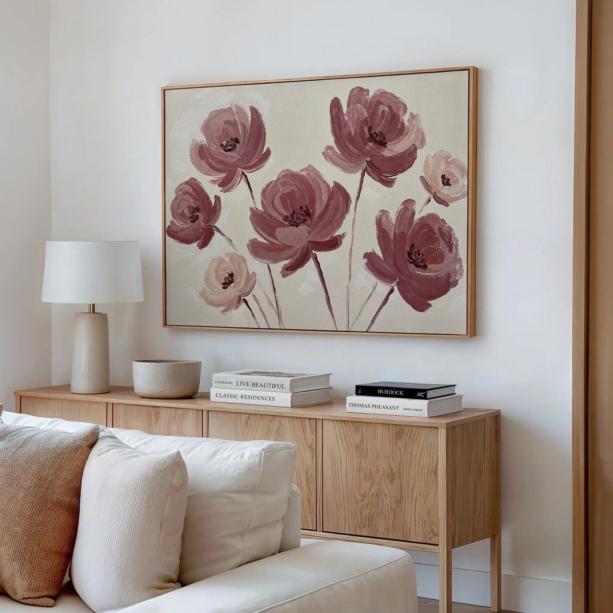 A modern living room with a light wood console, stacked books, a white table lamp, neutral decor, and the Autumn Rose Canvas Art featuring vintage-inspired pink and mauve flowers above a cream sofa on a white wall.