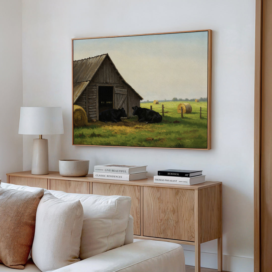 A cozy living room features a light sofa, wooden sideboard, and the Barnside Rest Canvas Art above—showing black cows resting by a barn in a rural field with hay bales.