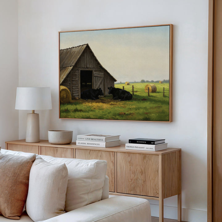 A cozy living room features a light sofa, wooden sideboard, and the Barnside Rest Canvas Art above—showing black cows resting by a barn in a rural field with hay bales.