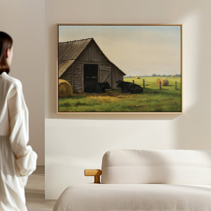 A person in white admires the Barnside Rest Canvas Art, featuring cows and a barn, displayed above a beige sofa and adding charm and warmth to the modern minimal living room.