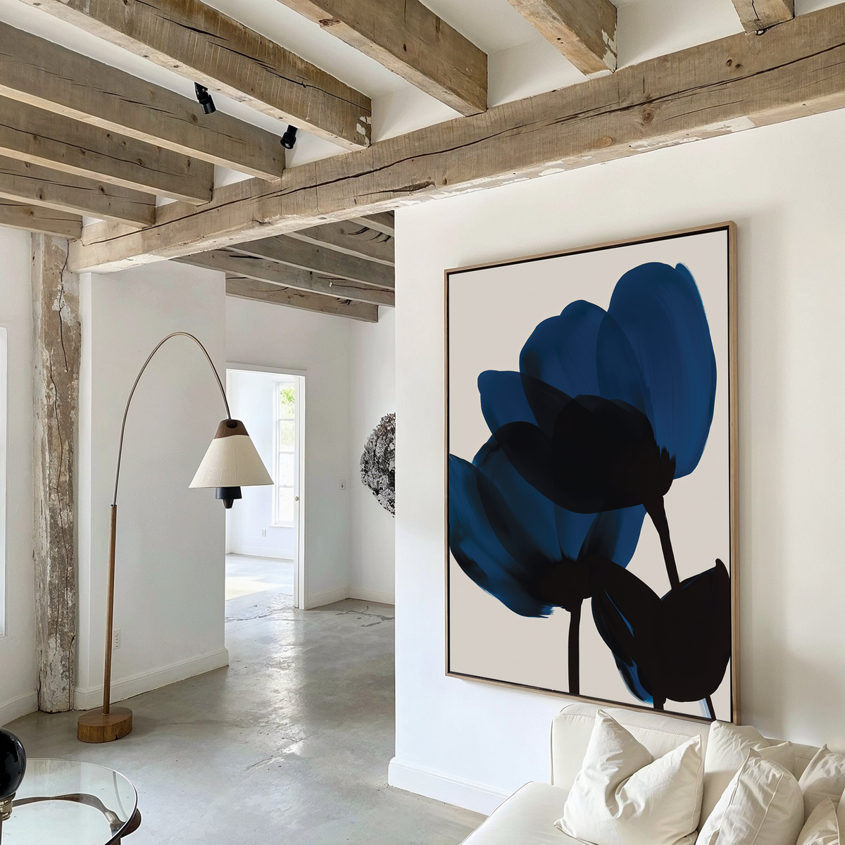 A modern living room featuring exposed wooden ceiling beams, a white sofa with a pillow, a tall curved floor lamp with a beige shade, and the Bloom Abstract Canvas Art displaying dark blue petals on the wall.