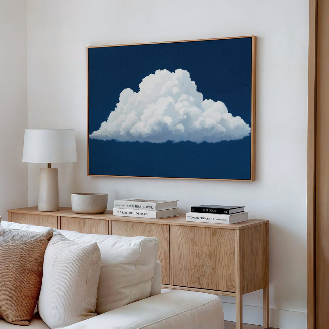 A modern living room features a light wood sideboard with books, a lamp, and a bowl, topped by the Blue Horizon Cloud Canvas Art—a minimalist piece depicting a fluffy white cloud on a dark blue background.