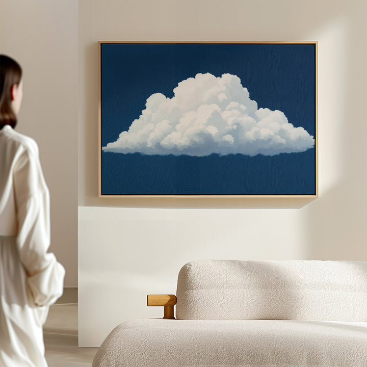 A person in a white shirt admires Blue Horizon Cloud Canvas Art—minimalist wall art with a fluffy cloud on blue—displayed above a modern beige sofa, ideal for elevating contemporary living rooms.