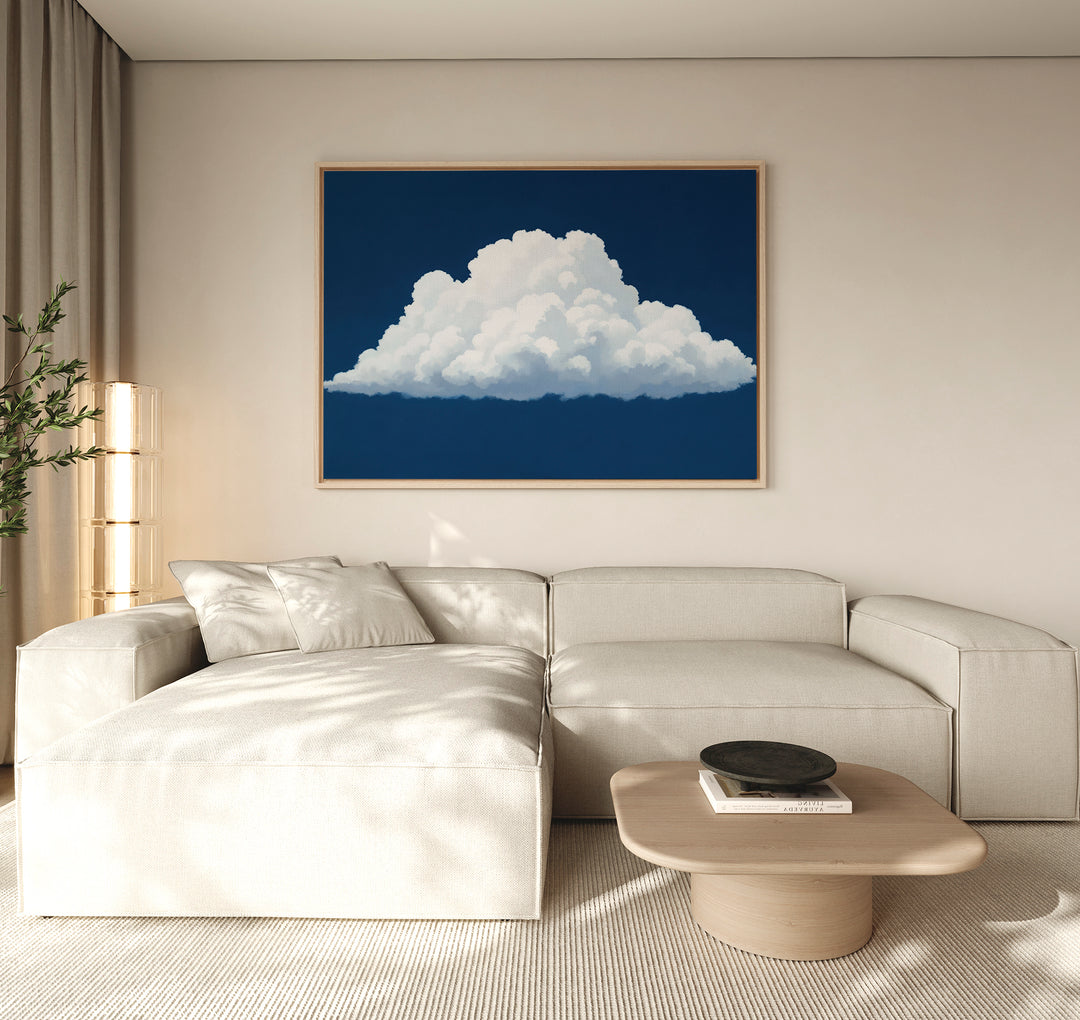 A modern living room with a beige sectional sofa, light wood coffee table, plant, and the Blue Horizon Cloud Canvas Art—a minimalist piece depicting a fluffy white cloud on a deep blue background—hanging above.