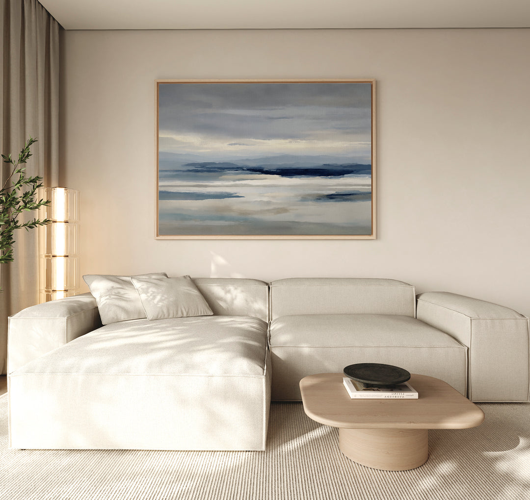 A modern living room with a light beige sectional sofa, low wooden coffee table, black bowl, tall plant, and minimalist wall art—a Blue Mist Horizon Canvas Art—serving as striking decor above the couch.