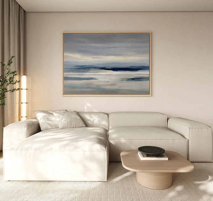 A modern living room with a light beige sectional sofa, low wooden coffee table, black bowl, tall plant, and minimalist wall art—a Blue Mist Horizon Canvas Art—serving as striking decor above the couch.