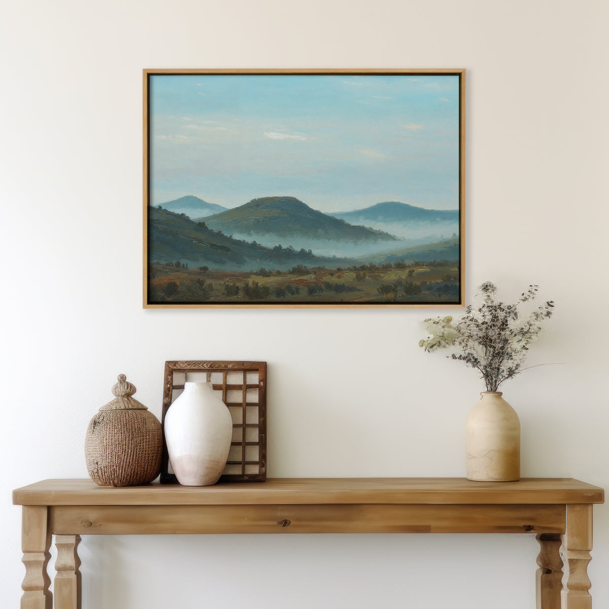 The Blue Ridge View Canvas Art, a framed landscape wall art of hills, hangs on a light wall above a wooden table with vases, a basket, dried flowers, and a small framed panel—perfect for creating serene bedroom decor.