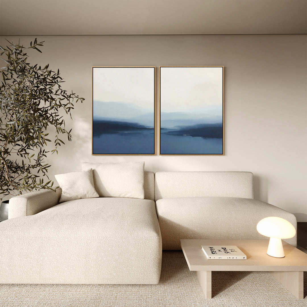 A modern living room with a cream sectional, small wood coffee table with lamp and book, minimalist decor, and the Blue Valley Mist Set of 2 wall art prints above the sofa. A tall plant stands in the corner.