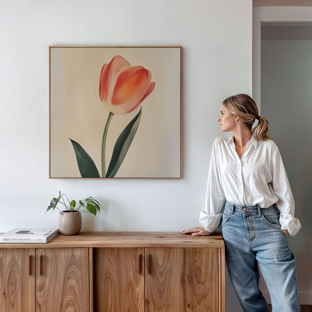 A woman in a white blouse and blue jeans leans against a wooden cabinet, admiring Blush Tulip Canvas Art; a potted plant and book on the cabinet add serene, minimalist vibes—ideal for bedroom decor.