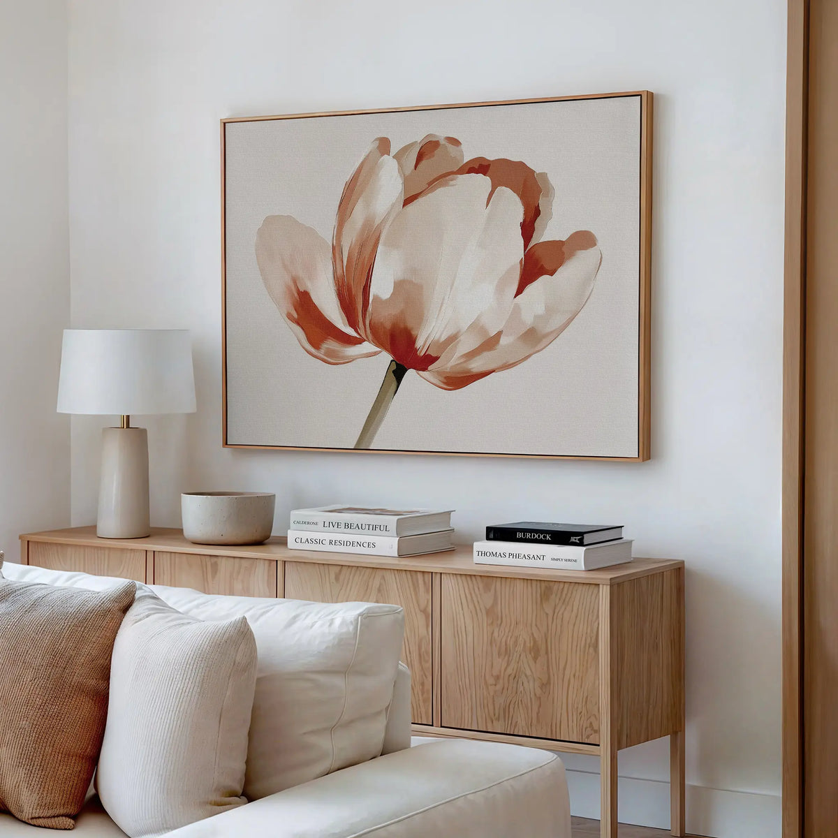 A modern living room with a light wood sideboard, white table lamp, decorative bowls, stacked books, and striking Blush Tulip Canvas Art featuring red and beige flowers above a cream-colored sofa.