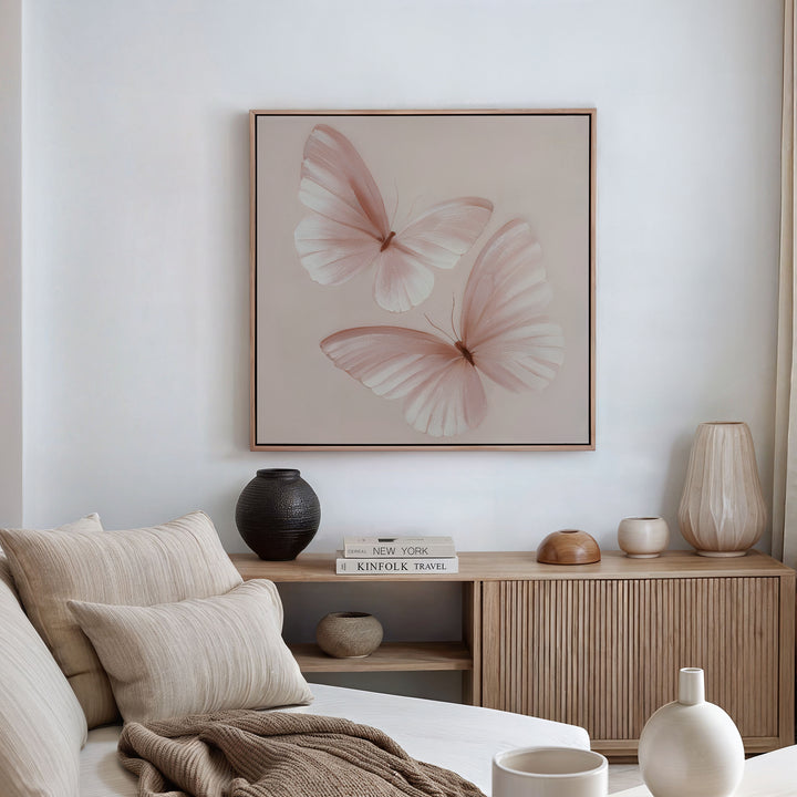 A cozy, neutral-toned living room with a beige sofa, soft cushions, a wooden sideboard, decorative vases, and blush pink wall decor featuring the Blush Wings Canvas Art above.
