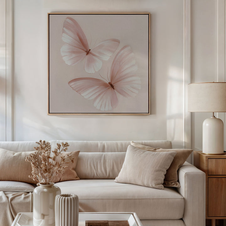 A bright, neutral-toned living room features a light sofa, beige pillows, and a vase of dried flowers. Above the sofa, the Blush Wings Canvas Art in soft blush pink adds charm. A table lamp rests on a wooden side table.