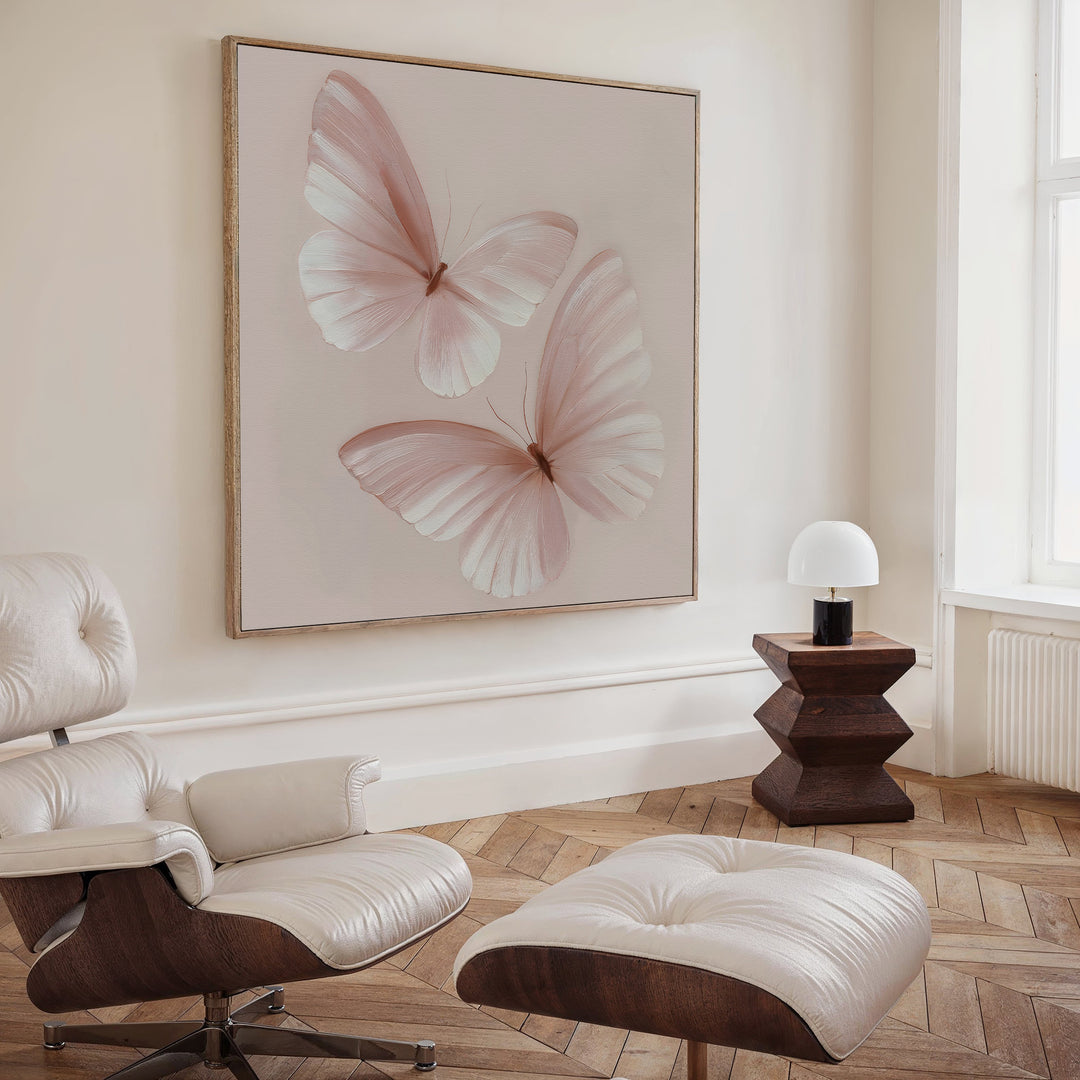 A modern living room features a white lounge chair and ottoman, a wooden side table with lamp, and the Blush Wings Canvas Art that brings soft pink flair. Light wood herringbone flooring and cream walls complete the relaxing space.