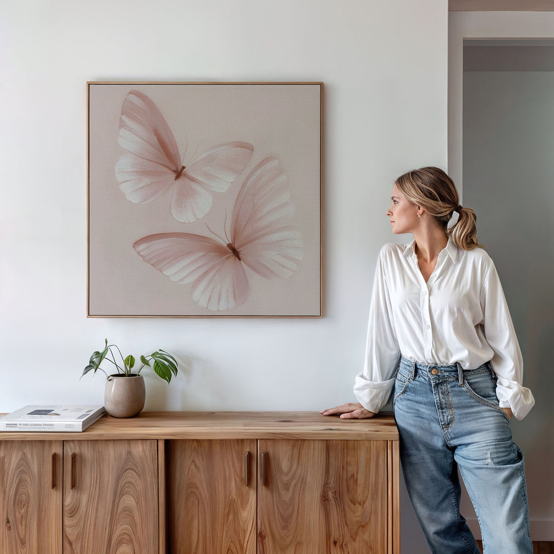 A woman in a white blouse and jeans stands by a wooden cabinet, admiring the Blush Wings Canvas Art with two light pink butterflies. A potted plant and book enhance the blush pink decor on the white wall.