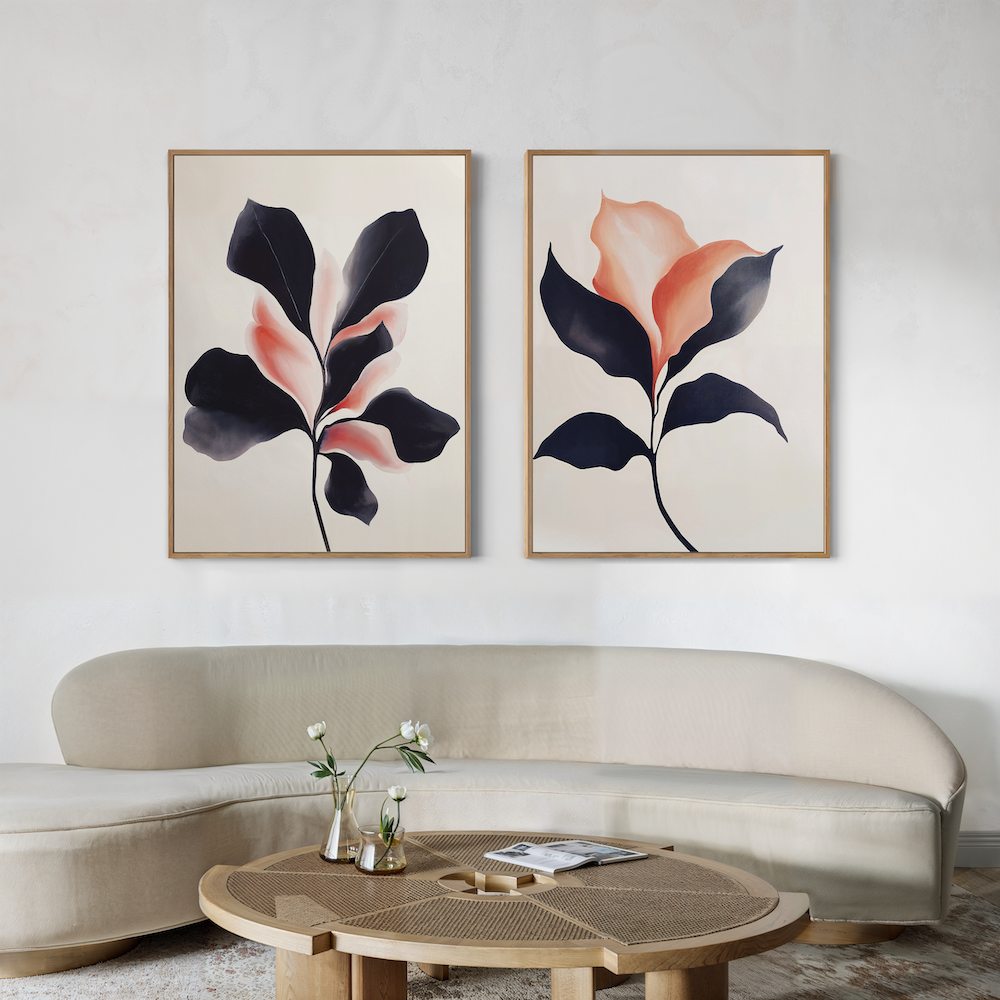 A modern living room featuring a curved beige sofa, a round wooden coffee table with vases and magazines, and the Blush & Shadow Set of 2 framed abstract floral artworks in black and peach tones on the wall.
