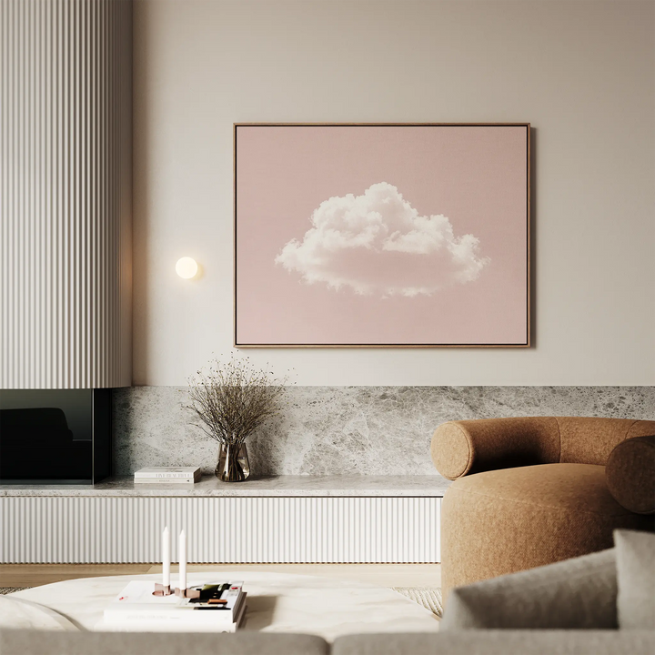 A modern living room with a beige curved sofa, marble accents, minimalist decor, a vase with dried flowers, and the Blushing Sky Canvas Art featuring a white cloud on a pink background hanging on the wall.