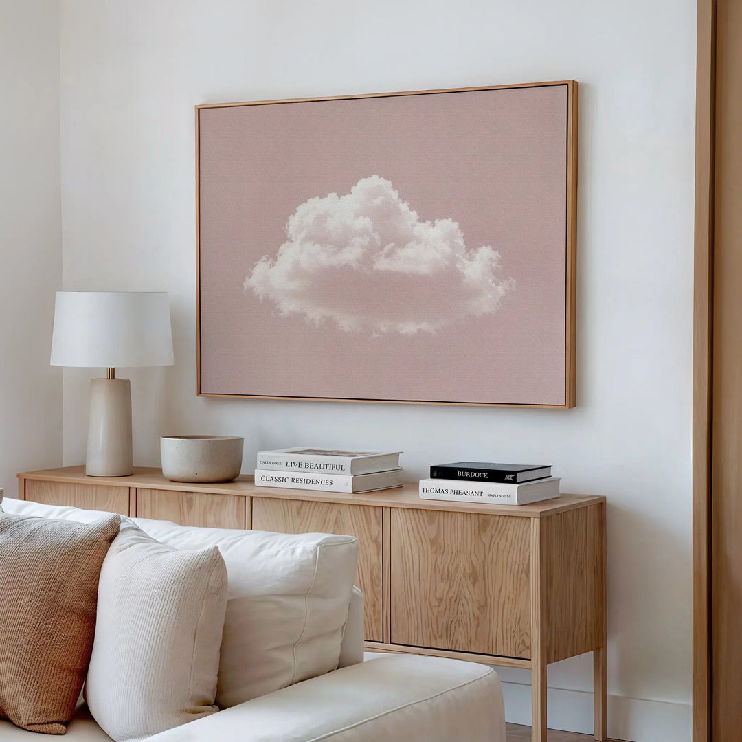 A modern living room with a beige sofa and light wood sideboard accented by books, a bowl, and a white lamp. Above the sideboard hangs the Blushing Sky Canvas Art, featuring a fluffy white cloud on a pale pink background.