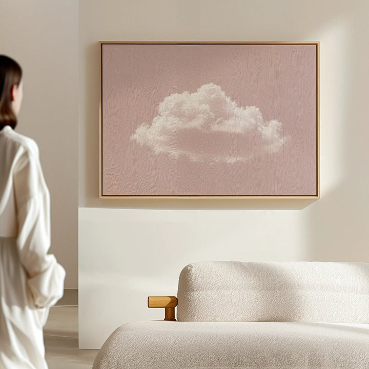 A person stands in a softly lit, minimalist room with a light sofa and the Blushing Sky Canvas Art—a framed print of a white cloud on pale pink—adding charm to the serene decor.