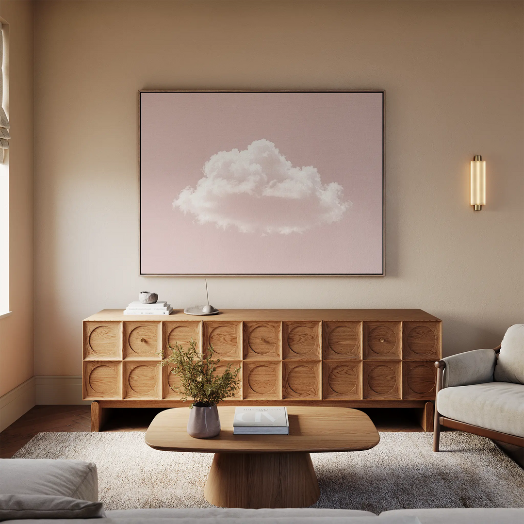 A cozy living room with a light wood sideboard, coffee table, and sofa features minimalist decor. The Blushing Sky Canvas Art, showing a fluffy white cloud on a pink background, hangs above the sideboard and complements the neutral tones.