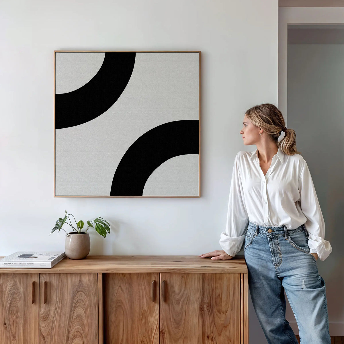 A woman in a white shirt and blue jeans stands by a wooden sideboard, admiring the Bold Orbit Canvas Art on the wall. A potted plant and books enhance the modern decor, creating a stylish yet inviting atmosphere.