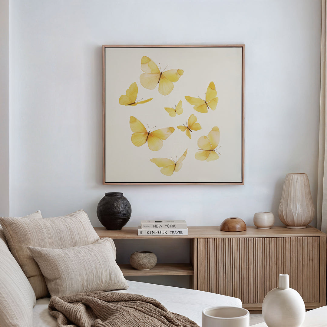 A minimalist living room features a beige sofa, neutral pillows, and a wooden sideboard. Above hangs the Butterfly Radiance Canvas Art, while decorative vases and books are neatly arranged on the sideboard.