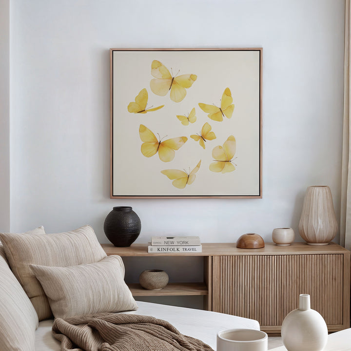 A minimalist living room features a beige sofa, neutral pillows, and a wooden sideboard. Above hangs the Butterfly Radiance Canvas Art, while decorative vases and books are neatly arranged on the sideboard.