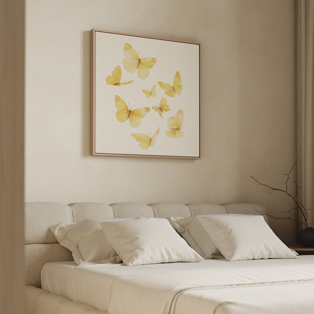A minimal bedroom features a beige upholstered bed with white bedding and the Butterfly Radiance Canvas Art of yellow butterflies above the headboard. A simple branch decor rests on the nightstand near the curtains.