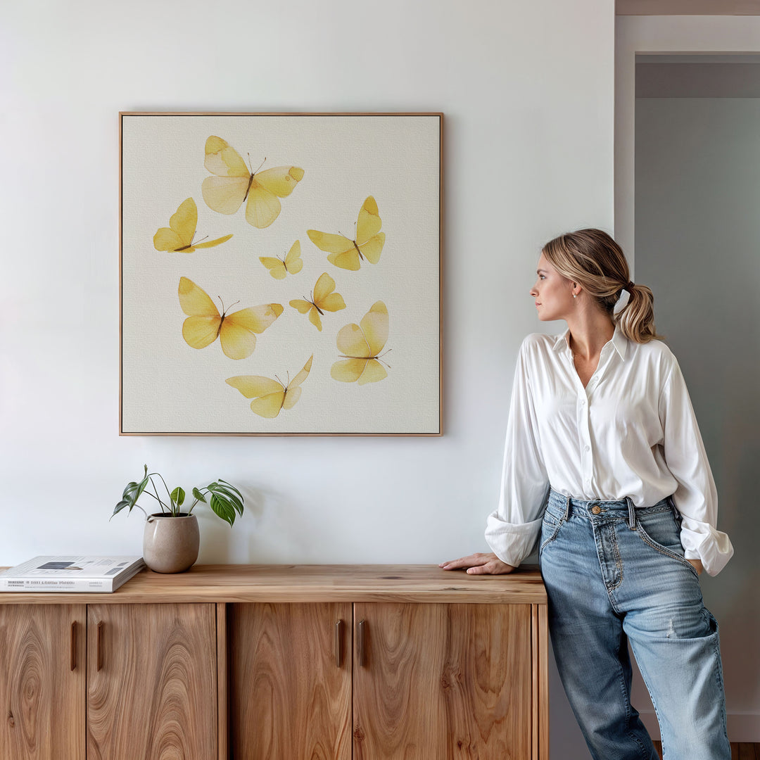 A woman in a white blouse and jeans leans against a wooden cabinet, admiring the Butterfly Radiance Canvas Art on a white wall. A potted plant and book atop the cabinet enhance the Nordic Park-inspired decor.