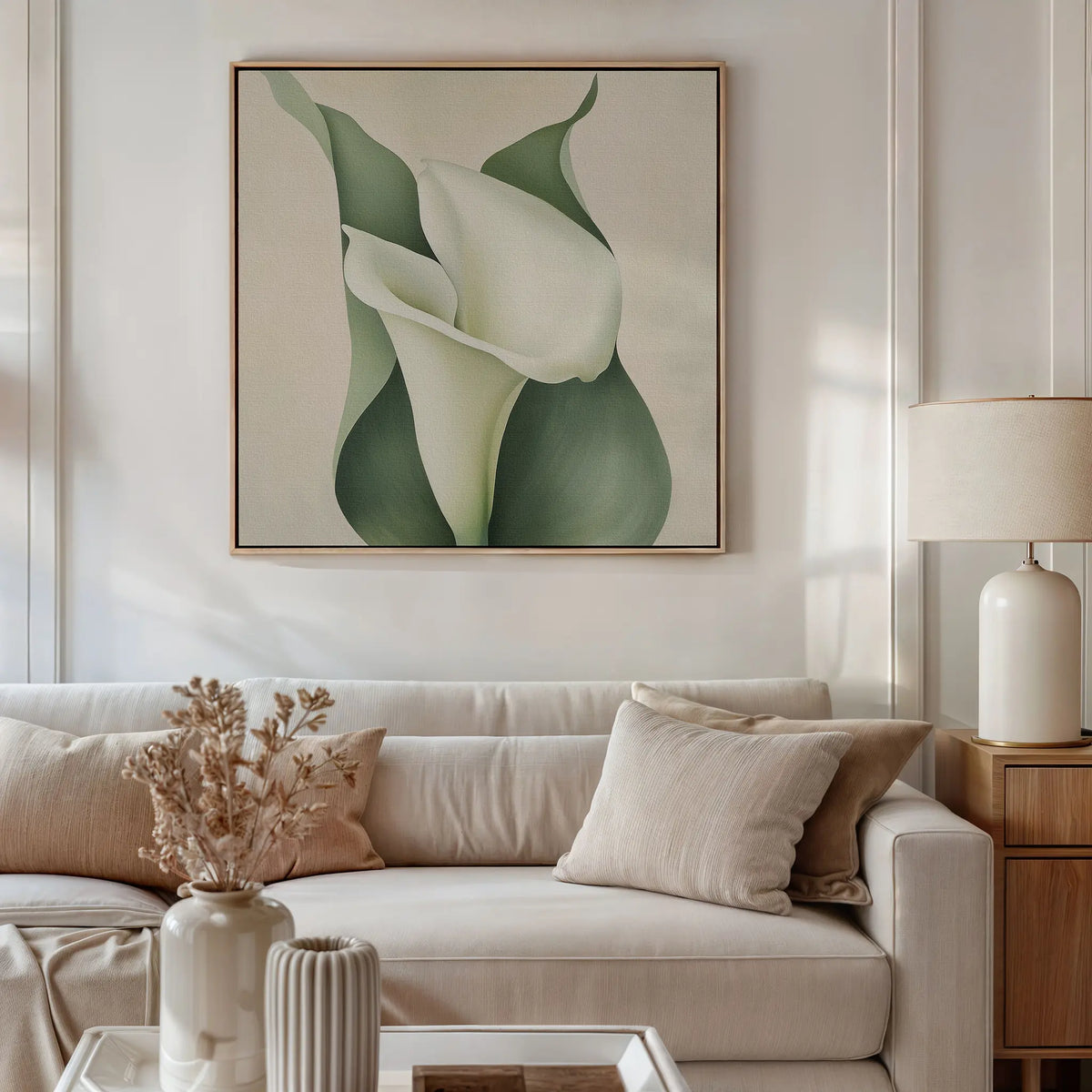 A neutral-toned living room with a light beige sofa, soft pillows, a side table and lamp, dried flowers in a vase, and striking floral wall art—the Calla Duo Canvas Art—featuring white calla lilies with green leaves as part of the modern decor.