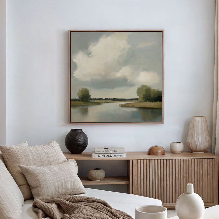 A cozy living room with a neutral sofa, beige pillows, a wooden sideboard with ceramics and books, and the Calm Passage Canvas Art depicting trees and a cloudy sky above a tranquil river.